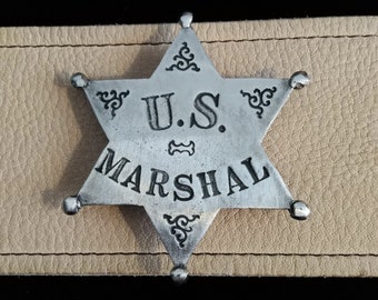 Könnte beinhalten: Ein silberfarbenes, sechszackiges Sternabzeichen mit dem Text "U.S. MARSHAL" in schwarzer Schrift. Das Abzeichen hat dekorative Verzierungen und ist vor einem hellbraunen Lederhintergrund angebracht. Das Abzeichen ist im Vintage-Stil.