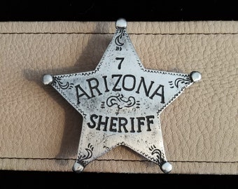 Puede incluir: Una placa de sheriff de estrella de cinco puntas de plata con las palabras "Arizona Sheriff" y el número "7" inscrito en ella.