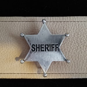 May include: A silver sheriff's star badge with five points and the word "SHERIFF" embossed on the front. The badge is attached to a piece of light brown leather.