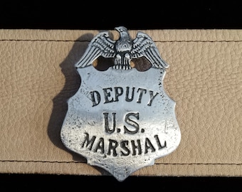 Puede incluir: Una insignia plateada de Deputy U.S. Marshal con un emblema de águila. La insignia en forma de escudo descansa sobre una superficie de cuero marrón claro. El texto 'DEPUTY U.S. MARSHAL' es visible.