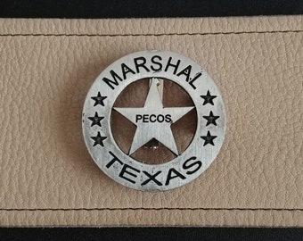 Puede incluir: Una insignia de metal plateado con una estrella de cinco puntas y las palabras "MARSHAL", "PECOS" y "TEXAS" alrededor de la estrella.
