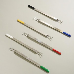 May include: A set of five stainless steel fondue forks, each featuring a long, slender metal shaft with two prongs at one end and a distinctively colored handle tip in black, blue, yellow, red, and green, arranged diagonally on a neutral surface.