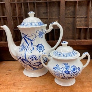 May include: A white teapot and sugar bowl with blue floral designs. The teapot has a tall, rounded body and a spout on the side. The sugar bowl has a lid and a handle on each side.