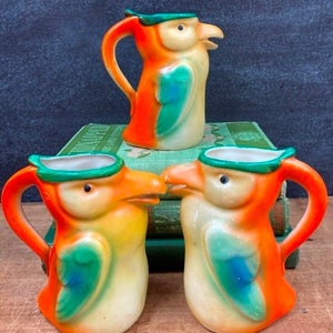 May include: Three ceramic pitchers shaped like parrots with green, orange, and white paint. The pitchers have a green beak and a green and blue wing design.