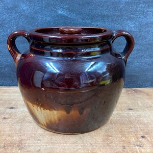 May include: A brown ceramic crock with a lid and two handles. The crock has a glossy finish with a gradient of colors, transitioning from dark brown to a lighter tan at the base. The lid has a small knob.