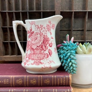 May include: A white ceramic pitcher with a red floral design, featuring a handle and spout. The pitcher is placed on top of two maroon books. A small succulent plant in a white pot is to the right.
