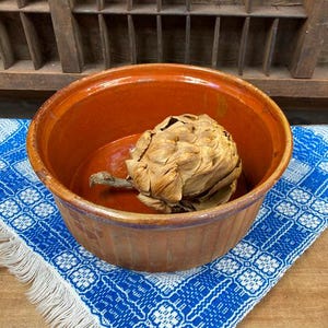 May include: A terracotta colored ceramic bowl with a dried artichoke inside. The bowl is sitting on a blue and white patterned fabric with white fringe.