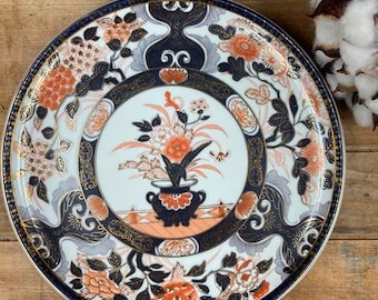May include: A white ceramic plate with a blue and gold rim. The plate features a floral design with orange, red, and blue flowers. The center of the plate has a vase with flowers in it.