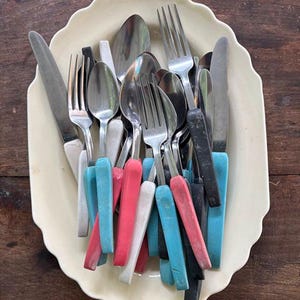 May include: A collection of stainless steel forks, spoons, and knives with colorful plastic handles in shades of blue, pink, white, and black, arranged inside an oval, cream-colored scalloped serving platter with a smooth, glossy finish.