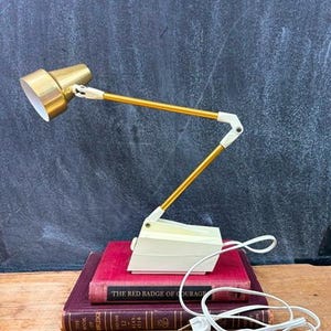 May include: A vintage desk lamp with a gold-colored shade and arm, and a cream-colored base. The lamp is positioned on top of two red books. A white power cord is visible.