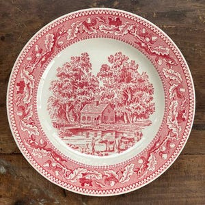 May include: A round ceramic plate featuring a red and white transferware pattern. The center depicts a rural scene with a house, trees, and cattle near a pond. The rim is decorated with a repeating border of oak leaves and acorns in red.