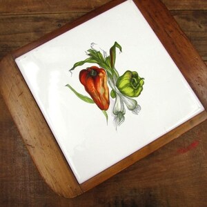 May include: A white ceramic trivet with a wooden frame. The trivet features a hand-painted design of a red pepper, a green pepper, and a bunch of green onions.