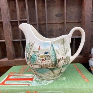May include: A white ceramic creamer with a winter scene painted on it. The scene features a small village with houses, trees, and a frozen lake. The creamer has a handle and a spout.