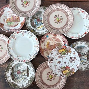 May include: A collection of antique decorative plates with various patterns. Some plates feature castle scenes, fruit designs, and winter landscapes. The plates have scalloped edges and are arranged on a dark wooden surface.