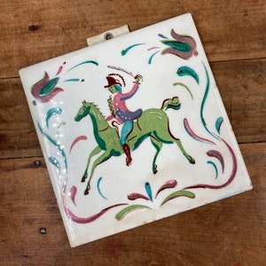 May include: A white ceramic tile with a hand-painted design of a person riding a horse. The tile has a green, pink, and blue floral border.