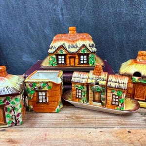 May include: A collection of ceramic cottage-shaped kitchenware. The set includes a sugar bowl, creamer, salt and pepper shakers, and a covered dish. The pieces are painted in orange, brown, and green, with thatched roofs and brick detailing.