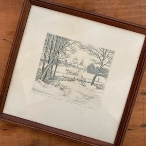 May include: A framed black and white print of a winter landscape scene. The artwork depicts trees, a village, and figures in a snowy setting. The frame is a dark brown wood. The print has a vintage aesthetic.