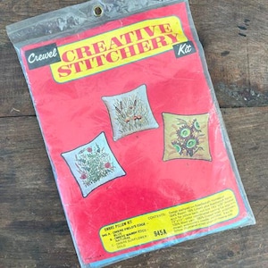 May include: A vintage "Creative Stitchery Kit" in a clear plastic bag. The red package features three embroidered pillow designs: wildflowers, cattails, and sunflowers. The kit includes the text "Ombre Pillow Kit" and the number "945A".