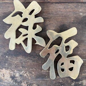 May include: Two brass-colored Chinese characters, possibly wall decor, rest on a weathered wooden surface. The characters are stylized and have cut-out details. The brass has a slight sheen, reflecting the light.