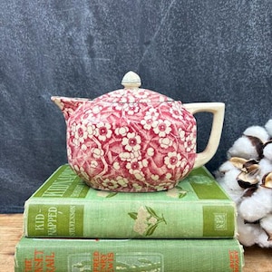 May include: A vintage teapot with a red and white floral pattern, resting on two antique green books. The teapot has a cream-colored handle and spout. The books are stacked on top of each other.
