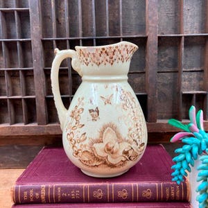 May include: A vintage cream-colored pitcher with a brown floral design and a butterfly. The pitcher is sitting on top of two burgundy books with gold lettering.