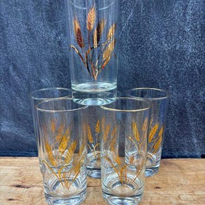 May include: Six clear highball glasses with gold wheat stalk designs and gold rims. The glasses are arranged on a wooden surface against a dark blue background. The wheat design is a prominent feature.