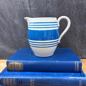 May include: A white and blue striped ceramic pitcher with a handle, sitting on top of two blue books. The pitcher has a spout for pouring. The books have gold lettering.