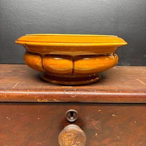 May include: A round, orange-glazed ceramic bowl with a scalloped design. The bowl sits on a wooden surface, and the background is a dark gray color. The bowl is a decorative piece.