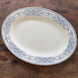 May include: An oval-shaped ceramic serving platter with a cream-colored center and a wide border featuring a repeating grey pattern of intricate floral motifs, geometric lattice panels, and delicate scrollwork along the outer rim edge.