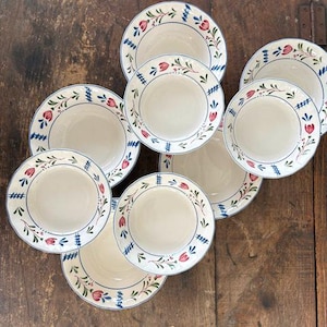 May include: A collection of white ceramic bowls with a floral and blue design. The bowls have a cream-colored interior and a decorative border featuring red flowers and blue accents. The bowls are arranged on a dark wooden surface.