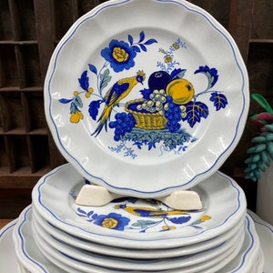 May include: A stack of white ceramic dinner plates with a blue and yellow floral design featuring a bird and fruit in a basket.