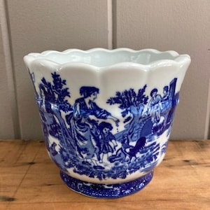 May include: A white ceramic planter with a blue and white floral design. The planter has a scalloped rim and a wide base.