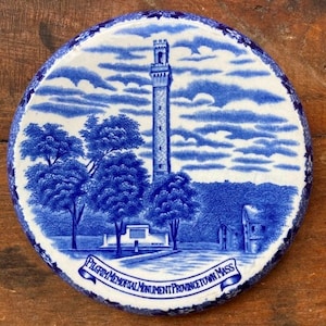May include: Blue and white ceramic plate with a depiction of the Pilgrim Monument in Provincetown, Massachusetts. The plate features a blue and white illustration of the monument, trees, and a cloudy sky. The text "Pilgrim Memorial Monument Provincetown Mass" is written in a blue banner at the bottom of the plate.