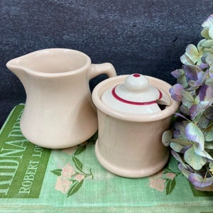May include: A cream-colored ceramic creamer pitcher with a curved handle and a matching lidded sugar bowl featuring a small knob handle and a thin red circular accent line on the lid, both resting on a green book with floral patterns.