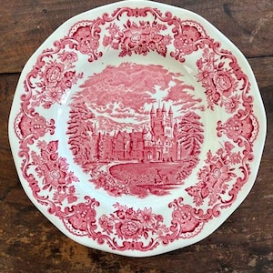 May include: A round, white ceramic plate with a red floral border and a central scene of a castle and landscape. The plate has a scalloped edge and is set on a wooden surface.