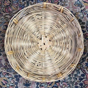 May include: A round, woven wicker basket with a natural brown color. The basket is made of thin, tightly woven strips of wicker, creating a smooth, even surface. The basket has a circular shape with a slightly raised rim.