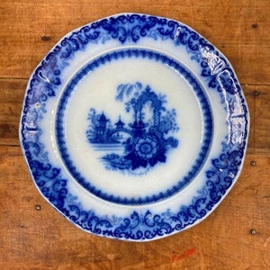 May include: A round, antique white plate with a blue floral and landscape design. The plate features a detailed blue border and a central image of a bridge and floral arrangement. The plate is set against a wooden surface.