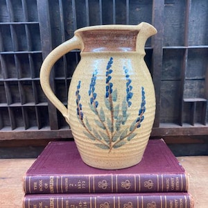 May include: A yellow ceramic pitcher with a brown rim and a floral design of blue and green lavender flowers. The pitcher has a handle and is sitting on top of two burgundy books.