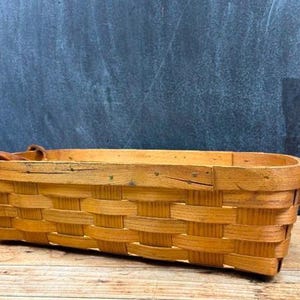 May include: A rectangular woven wood basket with a light brown finish, featuring a flat base, vertical side walls, and two brown leather handles attached to the sides with metal rivets. The basket has a visible wood grain and a woven pattern.