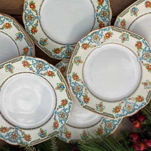May include: A set of six white ceramic dinner plates with a decorative floral pattern in shades of blue, green, orange, and gold. The plates have a scalloped edge and a raised rim.