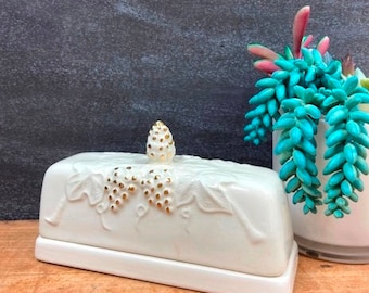 May include: White ceramic butter dish with a gold leaf design of grapes and vines. The lid has a gold leaf pine cone finial.