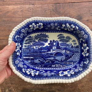 May include: A blue and white oval serving dish with a scalloped edge. The dish features a blue and white transferware design of a bridge, trees, and a river.