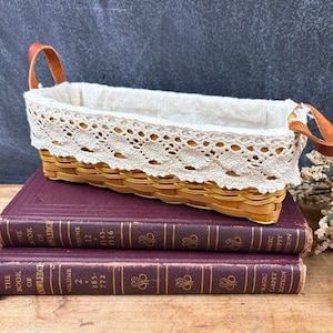 May include: A rectangular woven basket with light brown splints, featuring two tan leather handles on the ends. The basket is lined with off-white fabric and finished with a decorative cream-colored lace trim along the top edge.