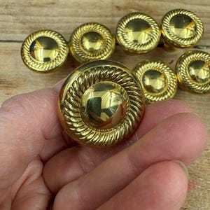 May include: A collection of eight gold-colored, round cabinet knobs. Each knob features a central, raised circle surrounded by a rope-like textured border. The knobs are displayed on a wooden surface.