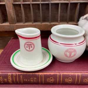 May include: Two vintage white ceramic creamer and sugar containers with red and green trim. The containers have a red and white logo with a snake and the letters "U.S."