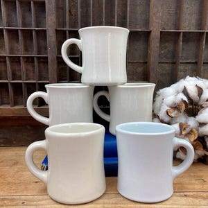 May include: Five white ceramic mugs with a rounded shape and a single handle. One mug is a lighter shade of blue.