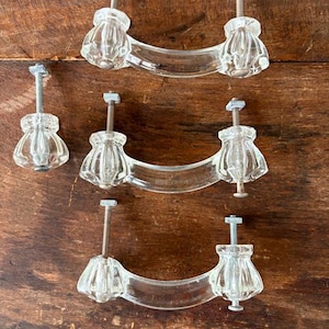May include: Clear glass cabinet pulls with a decorative, fluted design. The pulls have a curved handle and are attached with silver-colored screws. The image shows three pulls and one knob on a wooden surface.