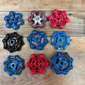 May include: Nine vintage water valve handles in various colors, including blue, red, and black. The handles are arranged on a weathered wooden surface, showcasing their aged appearance and unique designs. These are likely for industrial or rustic decor.