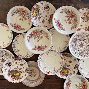 May include: A collection of vintage ceramic plates and saucers with floral and fruit patterns. The plates feature a cream-colored background with colorful flowers, fruits, and intricate designs. The arrangement includes various sizes and patterns, showcasing a classic aesthetic.