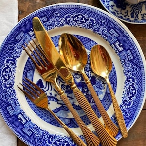 May include: Four gold-toned flatware pieces, including a knife, two forks, and a spoon, are arranged on a blue and white patterned plate. The plate has a white center and a blue border with a floral design.